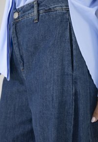 Donkerblauwe denim broek met een ontspannen pasvorm, voorzien van plooien aan de voorkant, een knoopsluiting en stiksel details. Gecombineerd met een lichtblauw overhemd.