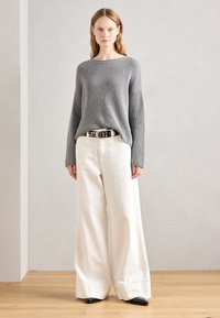 Grijze geribbelde trui met brede mouwen, gecombineerd met hoog-gewende witte wijde broek en een gemusterde zwarte riem. Zwarte puntige schoenen.