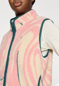 Γιλέκο απ ό φλις με ροζ και κρεμ σχεδιάγραμμα σπειροειδούς μορφής, πετρόλ φερμουάρ και τελειώματα. Διαθέτει ψηλό γιακά και αμάνικη κοπή, μαλακή υφή.