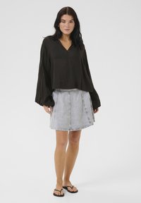 Schwarze, leichte Bluse mit langen Ärmeln und V-Ausschnitt, kombiniert mit einem hellen Denim-Rock mit A-Linien-Form und sichtbaren Nähten. Schwarze Flip-Flops.