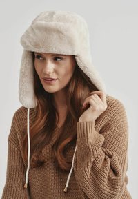 Sztuczny futrzany kapelusz w kremowym kolorze z klapkami na uszy, z białymi sznurkami i złotymi akcentami. Noszony z brązowym swetrem robionym na drutach.
