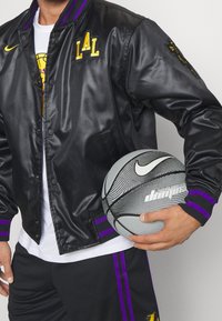 Chaqueta bomber de satén negra con detalles en púrpura y amarillo, con un logo en el pecho. Sosteniendo un balón de baloncesto gris con líneas negras y un logo.
