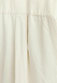 Tessuto crema chiaro con una texture a coste. Presenta pieghe e una cucitura, indicante un design di indumenti casual. Aspetto morbido e liscio.