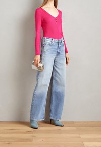 Frau trägt ein leuchtend pinkes Langarmoberteil, ausgewaschene blaue Jeans mit weitem Bein und blaue Stiefeletten mit Absatz, während sie eine silberne Clutch vor einem schlichten Hintergrund hält.