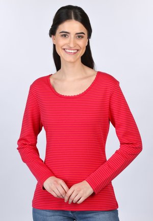 Rote und rosa gestreifte Langarmbluse mit Rundhalsausschnitt und welligem Saum, aus weichem Material. Das Model steht und lächelt.