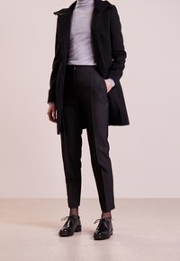 Sort uldfrakke over en grå højhalset trøje, kombineret med sorte skræddersyede bukser og sorte snøresko. Glatte teksturer og slank design.