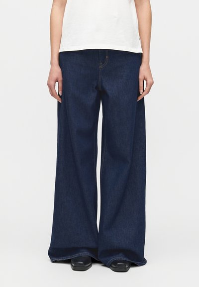 Personne portant un jean large en denim bleu foncé et des chaussures noires, debout devant un fond clair uni.