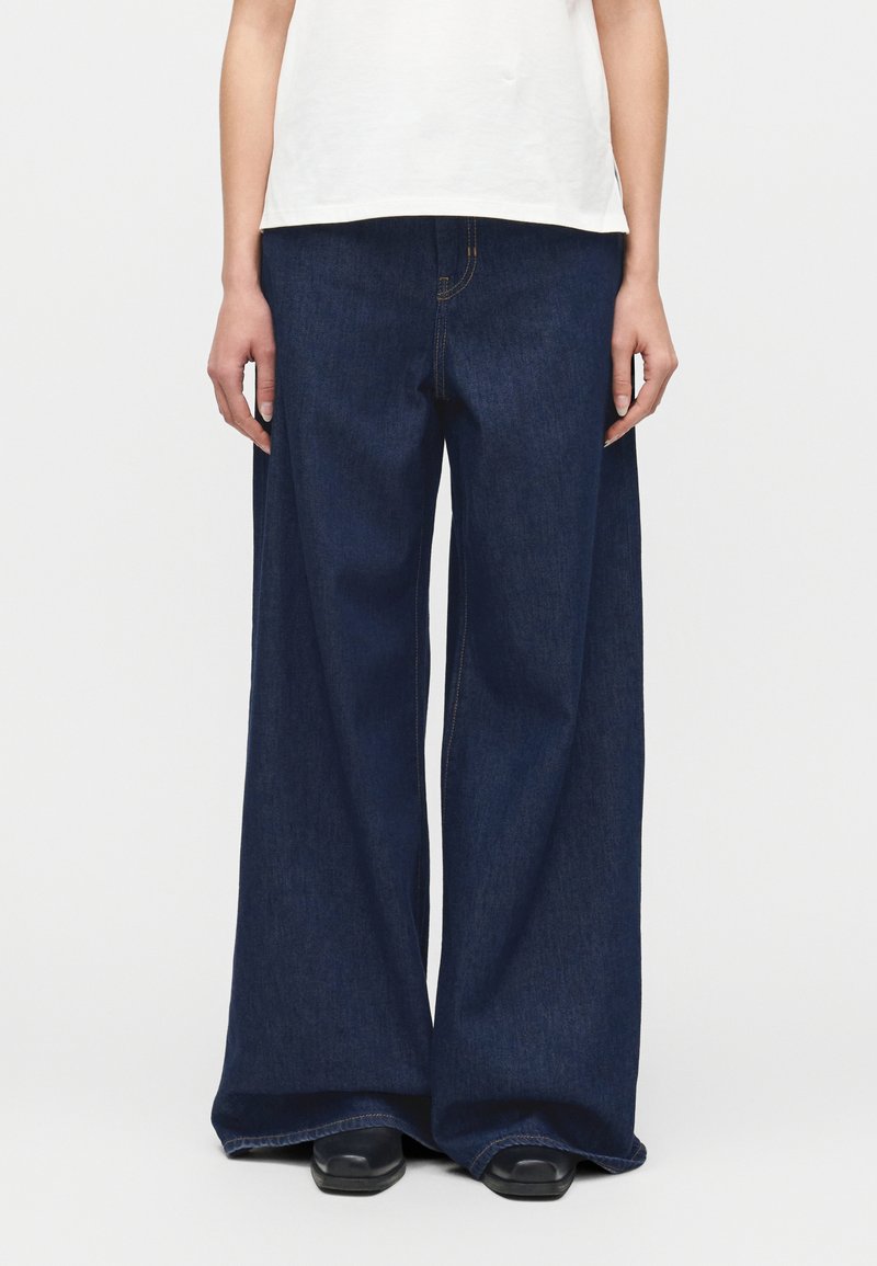 Άτομο που φοράει σκοτεινό μπλε τζι ν παντελόνι φαρδύ και μαύρα παπούτσια, στέκεται μπροστά σε απλό φωτεινό φόντο.