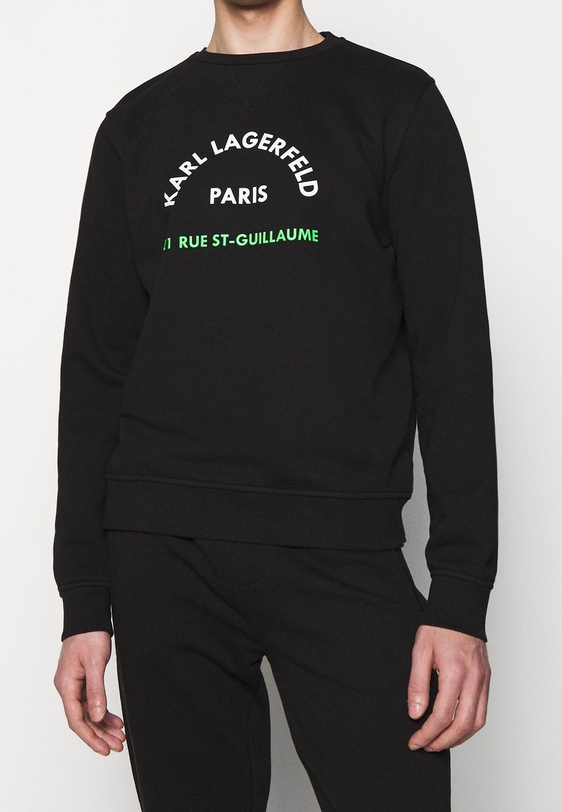 Sweat noir avec texte blanc et vert indiquant Karl Lagerfeld Paris et 21 Rue St-Guillaume, porté par une personne debout.