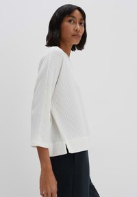 White ribbed sweater with 3/4 sleeves, featuring a wide hem and side slits, paired with dark trousers, against a plain background.