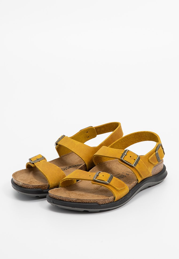 Sandales en daim jaune avec des brides croisées, des boucles en métal réglables et une semelle intérieure texturée marron sur une semelle en caoutchouc noire.