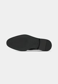 Sola de um sapato de vestir preto com uma superfície lisa e ligeiramente texturada e um salto baixo empilhado, contra um fundo branco.