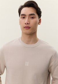 Mladý muž s krátkymi tmavými vlasmi, oblečený v svetlobéžovom svetri s okrúhlym výstrihom a malým bielym logom "II" na hrudi, neutrálne pozadie.