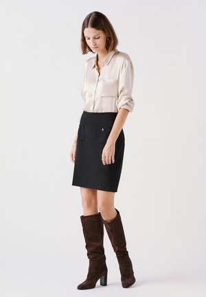Donna che indossa una blusa di raso color crema, una gonna nera con tasche frontali e stivali al ginocchio in camoscio marrone scuro, in piedi davanti a uno sfondo bianco.