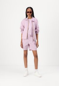 Lichtpaars jasje met elastische manchetten, bijpassend shirt en korte broek. Witte sneakers en rode zonnebril maken de look compleet. Glad stoftextuur.