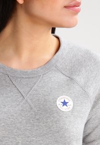 Grauer Hoodie aus strukturiertem Stoff, mit geripptem Rundhalsausschnitt und einem runden Converse All Star Logo, das einen blauen Stern und rote Akzente aufweist.