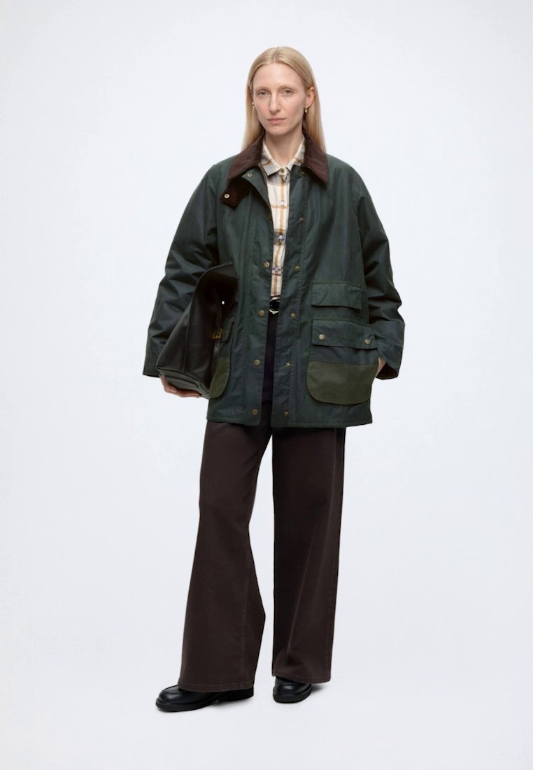 Femme blonde porte une veste oversize vert foncé avec plusieurs poches, une chemise à carreaux, un pantalon marron à larges jambes, des chaussures noires, tenant un sac noir.