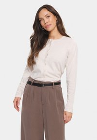 Svetlo bež bluza z dolgimi rokavi in gumbom na vratu, kombinirana s svetlo rjavimi hlačami z visokim pasom in zloženki ter črnim pasom.