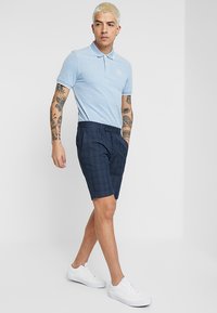 Lyseblå polo t-skjorte med korte ermer, kombinert med mørke rutete shorts. Brukt sammen med hvite joggesko, som viser en uformell, moderne stil.