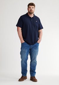 Marineblauwe polo met korte mouwen en een kraag, gecombineerd met lichtblauwe jeans en bruine schoenen. Opvallend logo op de borst.