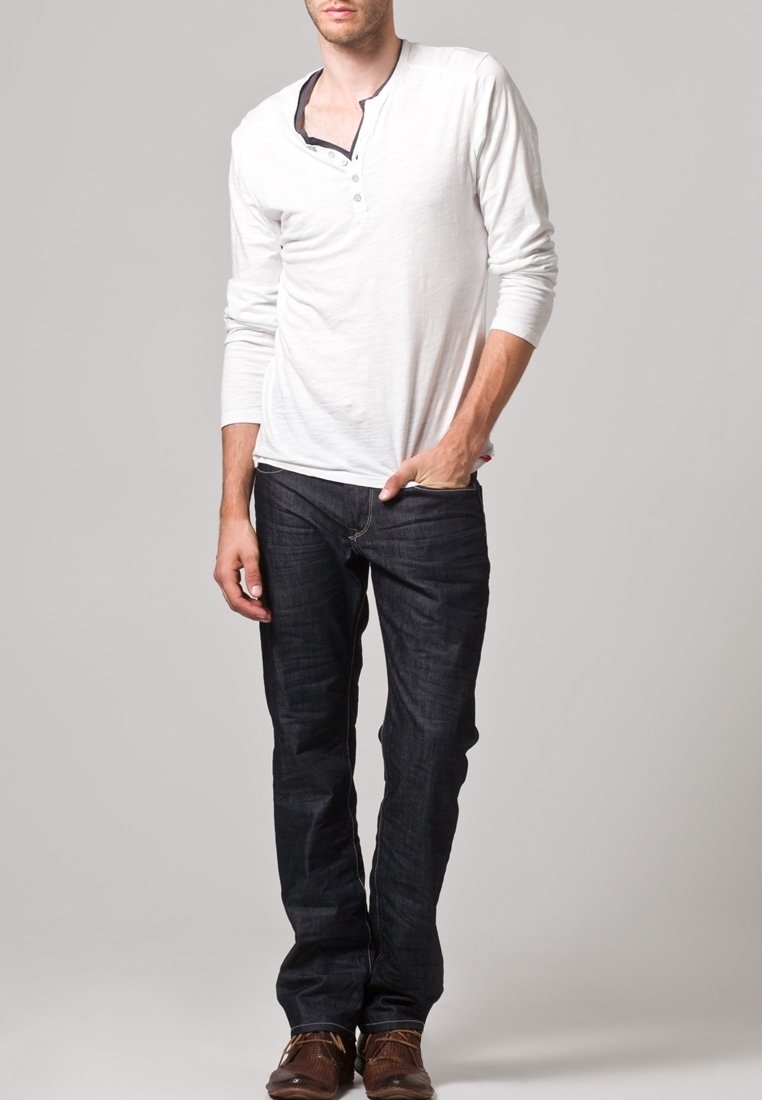 Camisa henley blanca de manga larga con tres botones, combinada con jeans de mezclilla oscuros. El conjunto incluye zapatos marrones y tiene un estilo informal.