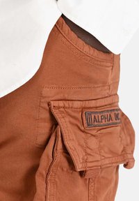 Close-up of brown cargo pants pocket with flap and "ALPHA" patch, worn with a white shirt tucked in.
