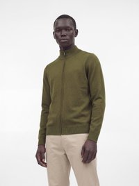 Homme debout portant un pull zippé vert olive et un pantalon beige sur un fond blanc uni.