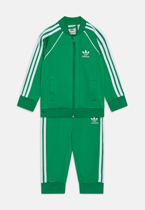 Zaļa Adidas sporta uzvalks ar baltām trīs svītrām. Jakai ir rāvējslēdzēja aizdare un ribota apkakle, bet biksēm ir elastīgas piedurknes.