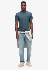 T-shirt blu heather a girocollo con un piccolo logo, abbinato a jeans azzurri chiari, una felpa grigia annodata in vita e sneakers beige.