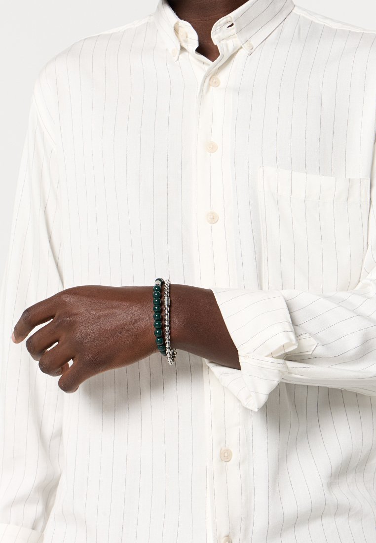 Chemise rayée blanche, boutonnée, avec une poche sur la poitrine. Le bracelet présente des perles vert foncé, des accents en argent et une chaîne argentée texturée.