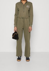 Combinaison vert olive avec un col zippé, une taille élastique et des manches longues. Associée à un sac à main en cuir texturé noir et des baskets sombres.