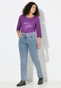 Kiharatukkainen nainen, jolla on päällään violetti pitkähihaisen paita, vaaleansiniset farkut violetilla vyöllä ja mustat nilkkurit, seisoo ja hymyilee.