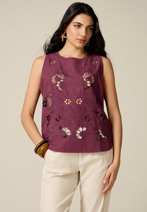 Mujer que lleva una blusa sin mangas color granate con bordado floral recortado, pantalones beige, pendientes de estrella de mar y pulseras color ámbar, posando sobre un fondo neutro.
