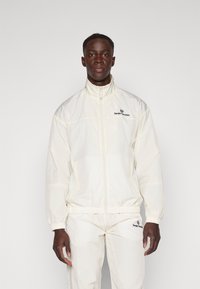 Hvit zip-up jakke laget av lett materiale med høy krage og elastiske mansjetter. Har et brodert logo på brystet.
