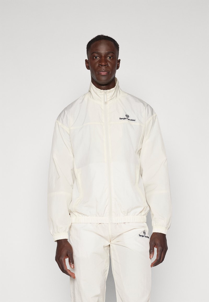 Hvit zip-up jakke laget av lett materiale med høy krage og elastiske mansjetter. Har et brodert logo på brystet.
