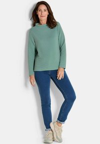 Groene geribbelde lange mouwen top gecombineerd met blauwe skinny jeans. Model draagt beige sneakers met mesh en camouflage accenten. Eenvoudige, casual uitstraling.