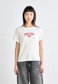 Camiseta blanca de canalé con el logo rojo de "TOMMY JEANS". Mangas cortas, cuello redondo y un corte relajado, combinada con pantalones de mezclilla grises.
