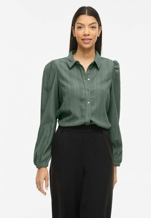 Kvinde iført en mørkegrøn struktureret knap-op bluse med lange ballonærmer og sorte bukser med høj talje, stående mod en ensfarvet baggrund.