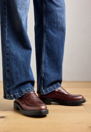 Persona che indossa mocassini in pelle marrone e jeans blu, in piedi su un pavimento di legno contro uno sfondo neutro.