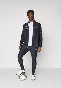 Svart sporty joggedress med hvite detaljer, som inkluderer en jakke med glidelås og innsnevrede bukser. Modellen står med en fotball og har på seg hvite joggesko.