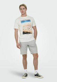 Bijela pamučna majica s grafikom šarenog plaže i natpisom "Sunshine State of Mind". Uparen s svijetlosivim šortsem i crnim cipelama.