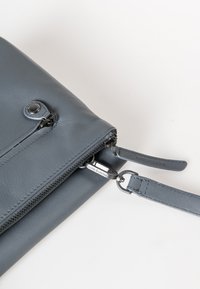 Graue Leder-Crossbody-Tasche mit einem Reißverschluss, ausgestattet mit einem runden Druckknopf und einem umgeprägten Trägerschild mit "aunts & uncles".