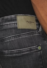 Černé džíny z denimu s zelenou koženou nášivkou s nápisem „Pepe Jeans London“. Materiál je hladký, s viditelným šitím a knoflíkem na přední kapse.