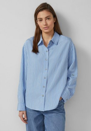 Jonge vrouw die een losse, lichtblauw-wit gestreepte blouse met knopen en een blauwe spijkerbroek draagt, staand tegen een effen achtergrond.