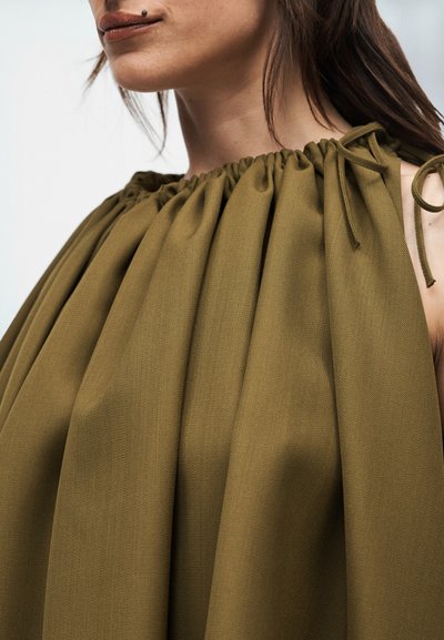 Κοντινό πλάνο γυναίκας που φοράει ένα αμάνικο φόρεμα ελιάς με μαζεμένο ύφασμα και δεμένα τιραντάκια στους ώμους.