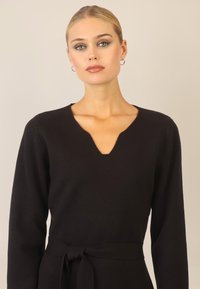 Schwarzes Strickkleid mit V-Ausschnitt, langen Ärmeln und einem gebundenen Taillendetail. Weiche Textur und taillierte Silhouette.