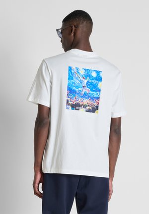 Man draagt een wit T-shirt met een kleurrijke afbeelding van een persoon die springt op een druk evenement onder een sterrenhemel op de rug.