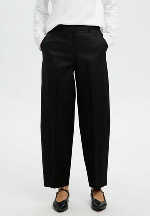 Pantalons larges taille haute noirs en tissu lisse avec plis sur le devant et passants de ceinture. Associés à des chaussures plates noires.