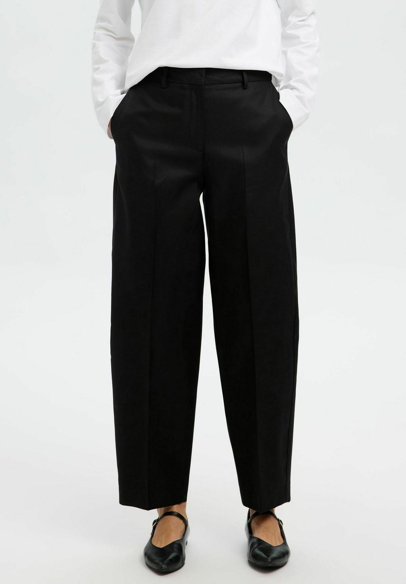 Zwarte high-waisted wijde broek gemaakt van gladde stof met voorplooien en riemlussen. Gecombineerd met zwarte platte schoenen.