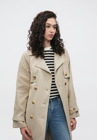 Trench-coat beige à double boutonnage, grands boutons jaunes et large col. Porté sur une chemise rayée et un jean bleu.